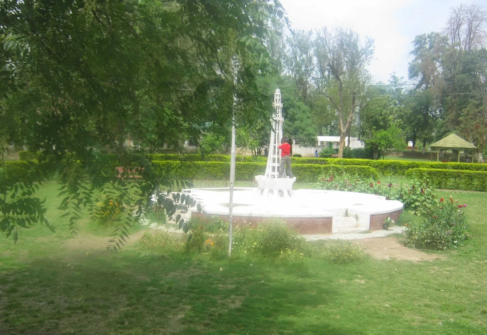 The white monument and fountain circle surrounded by hedged gardens, with a pagoda hut behind